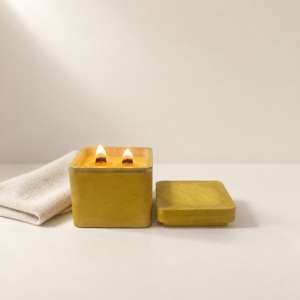 Sun Dried Linen luxury candle burning with two wooden wicks in a handcrafted yellow concrete vessel, with matching lid and folded linen.