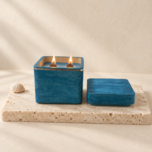 Lit Beach Walk luxury soy candle with two burning wood wicks on a travertine stone tile alongside a separate blue lid and small seashell.
