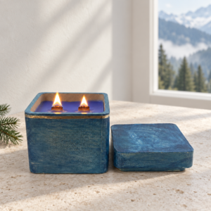 • An active burning Alpine Air luxury candle featuring dual wooden wicks lit with realistic flames inside an open blue concrete jar next to its lid on a textured stone surface.