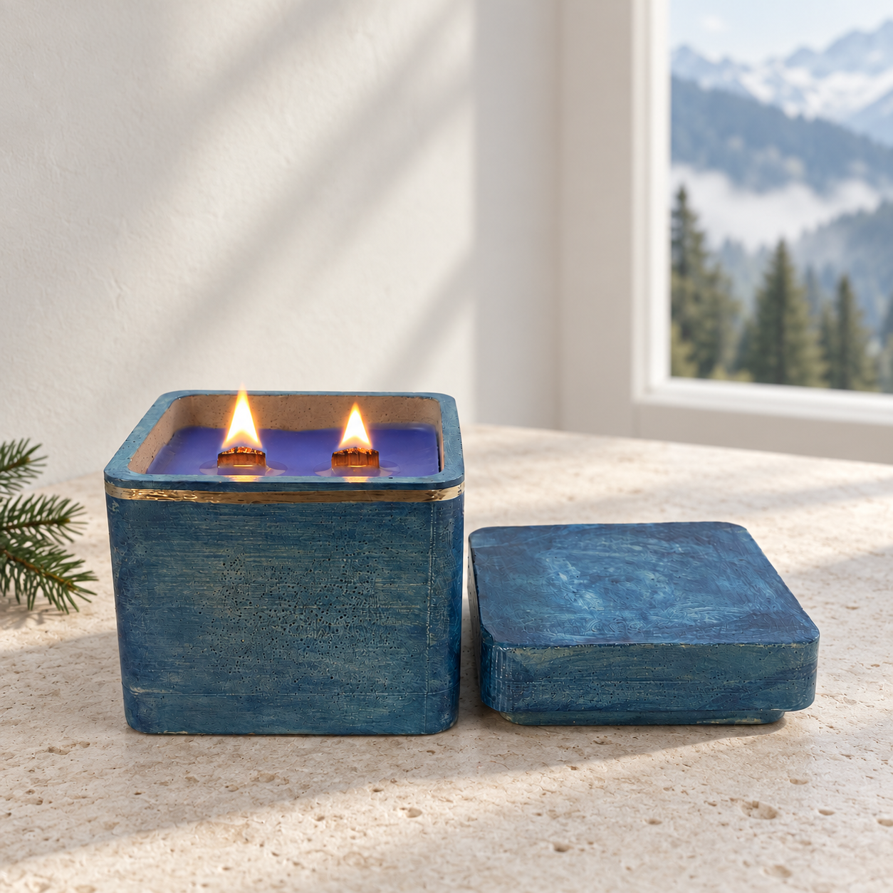 • An active burning Alpine Air luxury candle featuring dual wooden wicks lit with realistic flames inside an open blue concrete jar next to its lid on a textured stone surface.