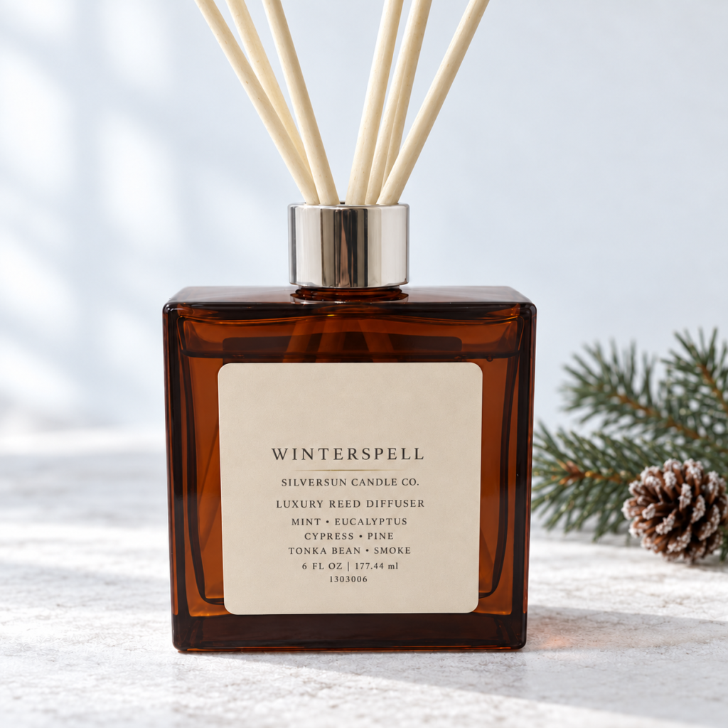 Winterspell luxury reed diffuser in an amber glass bottle with white reeds and silver collar, on a snow covered table.